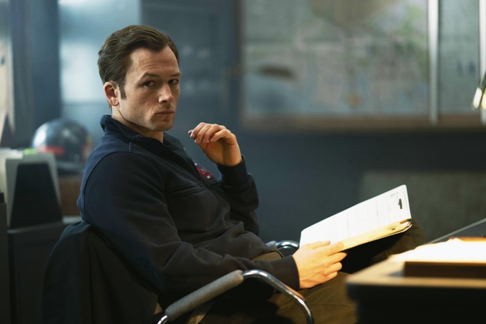 Taron Edgerton in Smoke, sat at a desk with a clipboard in his hands.