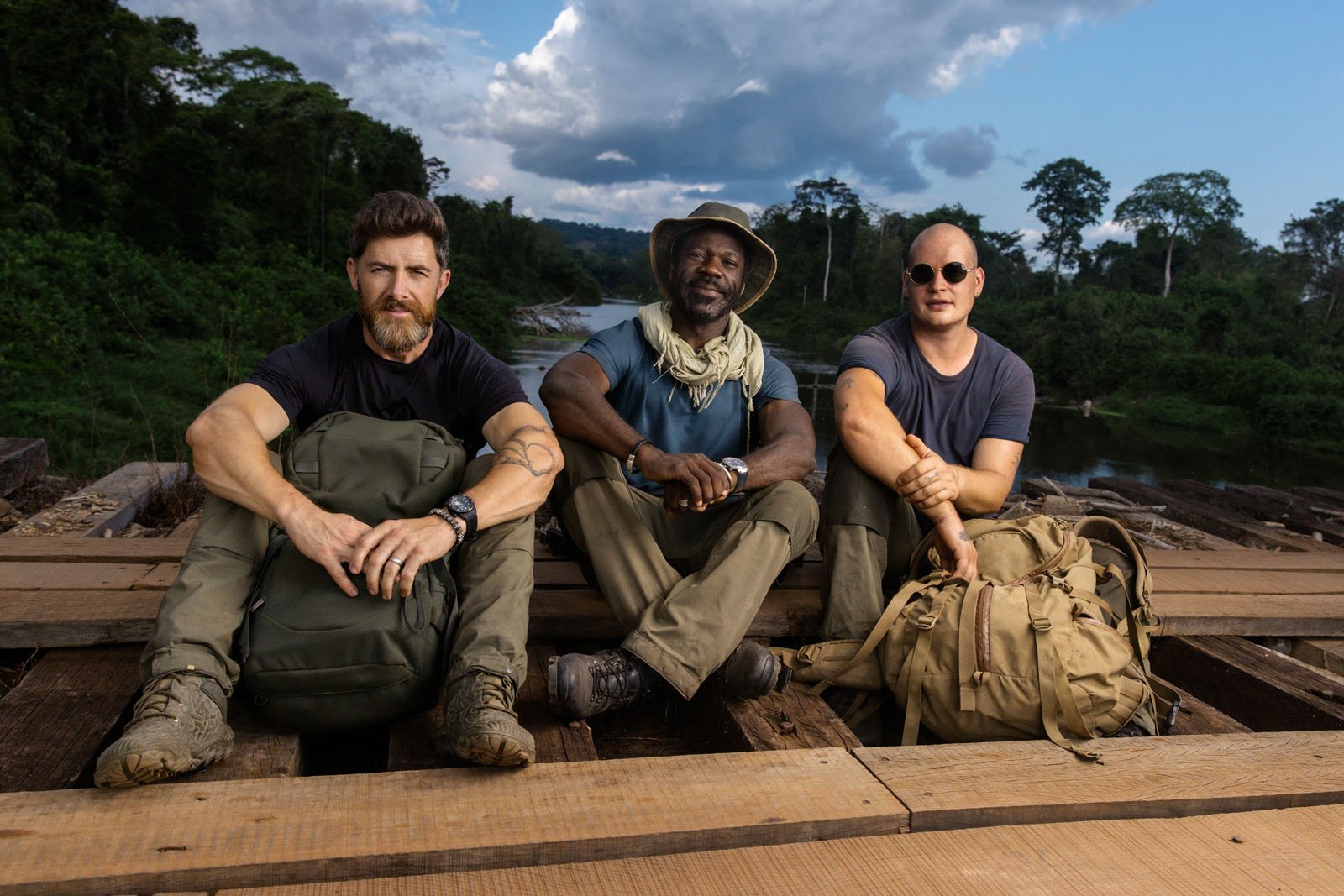Three men in outdoor gear sit on wooden planks in a lush, green forest setting with backpacks beside them. The sky is partly cloudy, and a river is visible in the background.