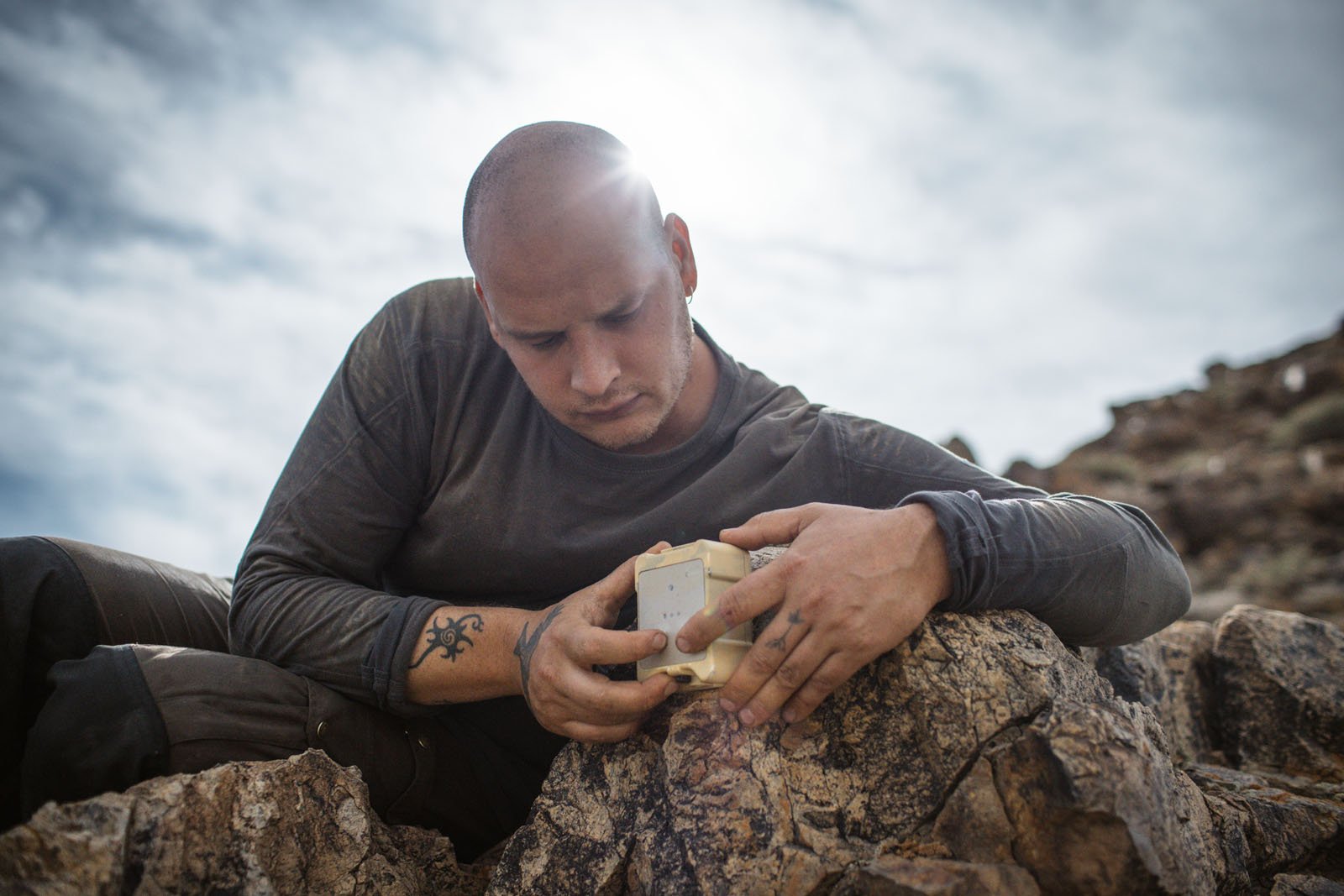 A person with a shaved head examines a small device while lying on rocks outdoors under a cloudy sky, with sunlight shining behind their head.