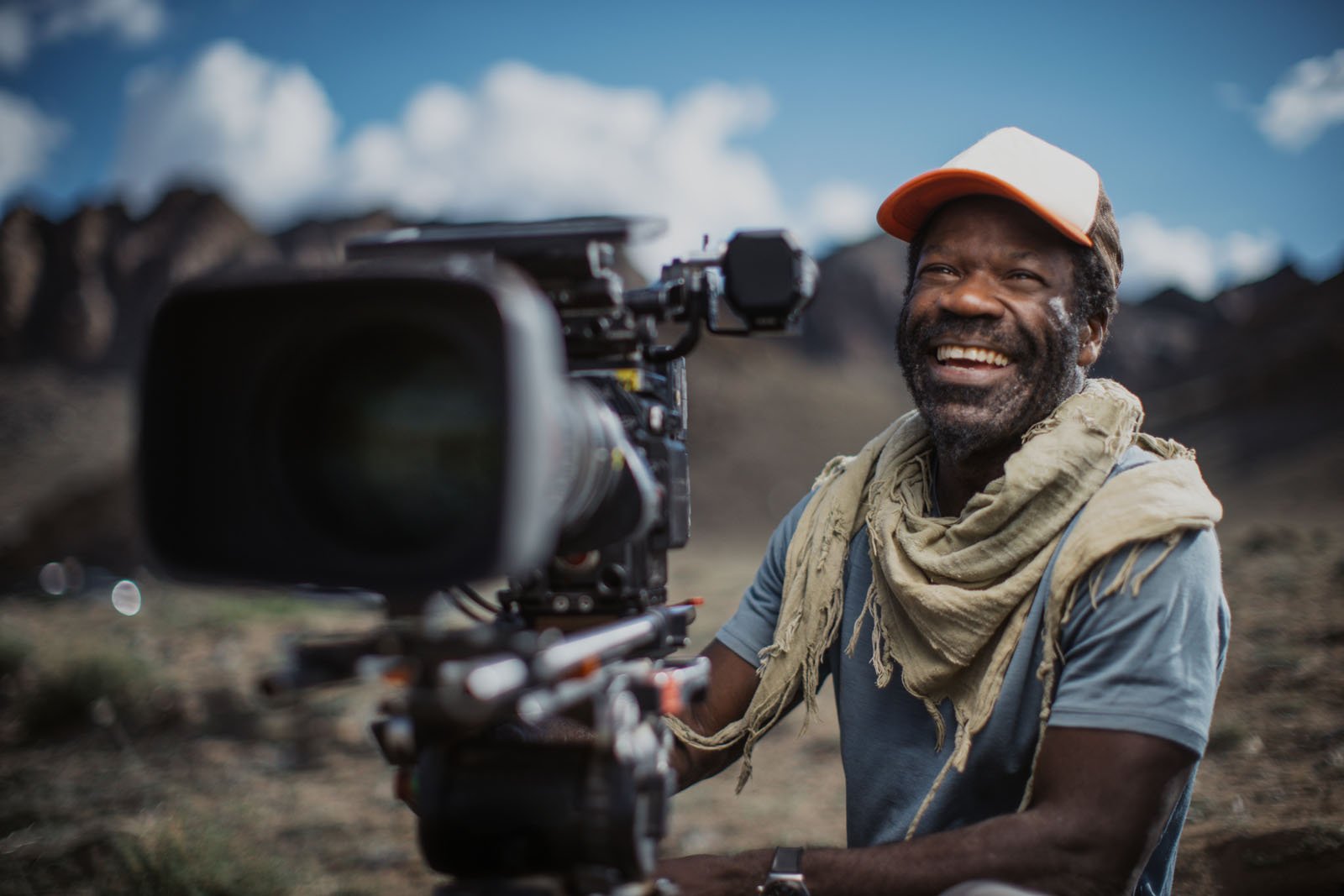 A smiling man wearing a cap and scarf sits outdoors beside a professional video camera, with mountains and a blue sky in the background.