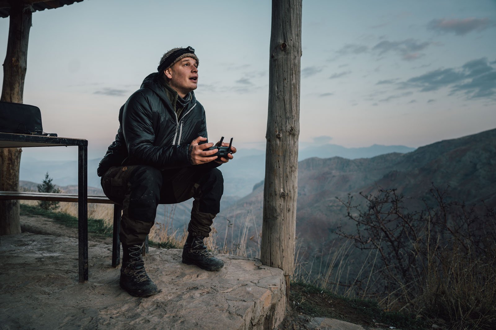 A person in outdoor gear sits on a stone platform under a wooden shelter, holding a remote control and looking up, with mountains and a cloudy sky in the background.