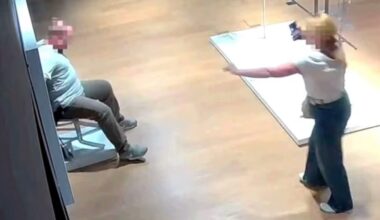 A security camera image shows a person sitting on a chair while another person stands nearby pointing a phone at them. Both faces are blurred, and the scene takes place in a well-lit indoor space with wooden floors.