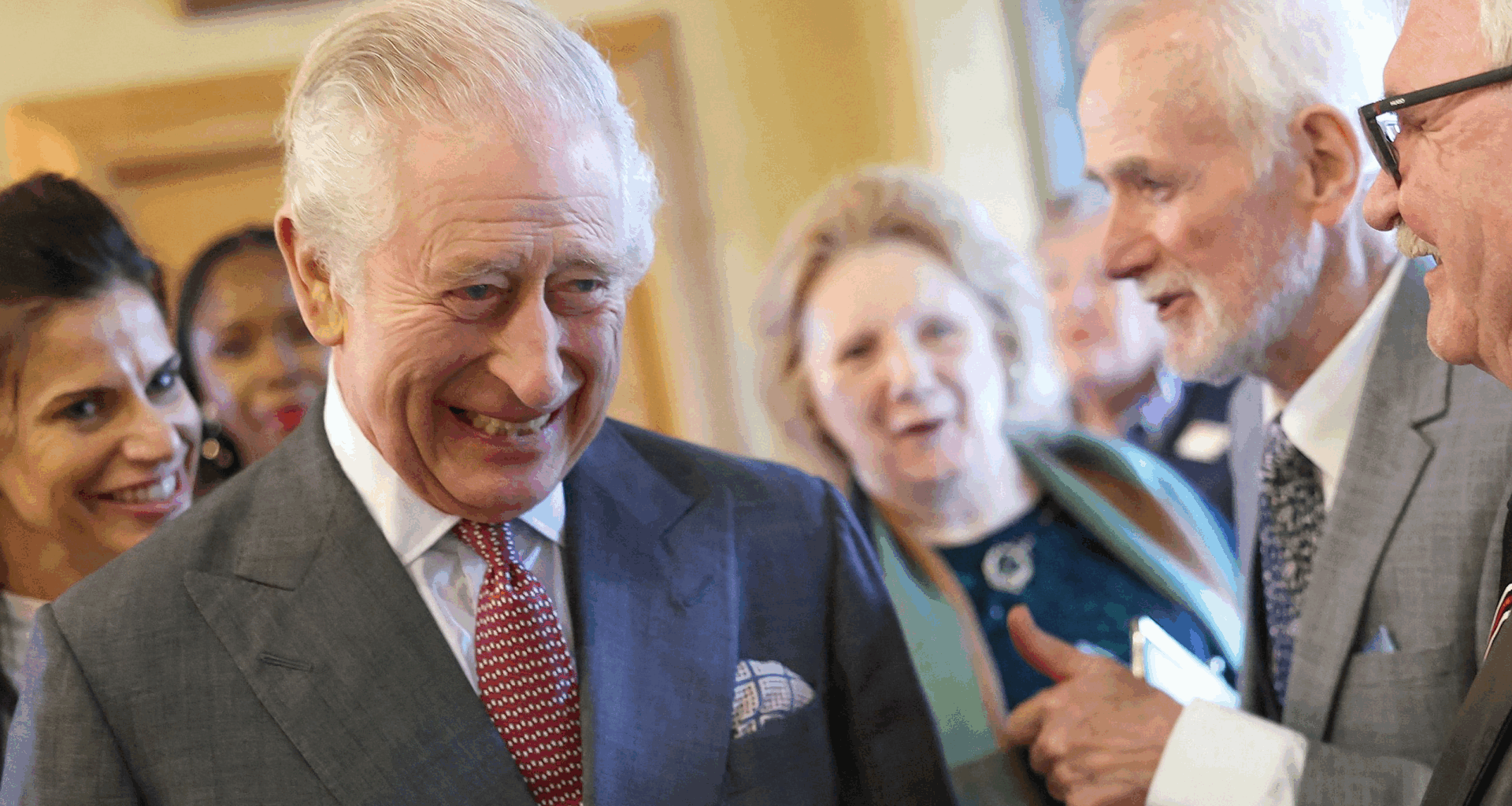 King Charles laughing in a gray suit and red tie