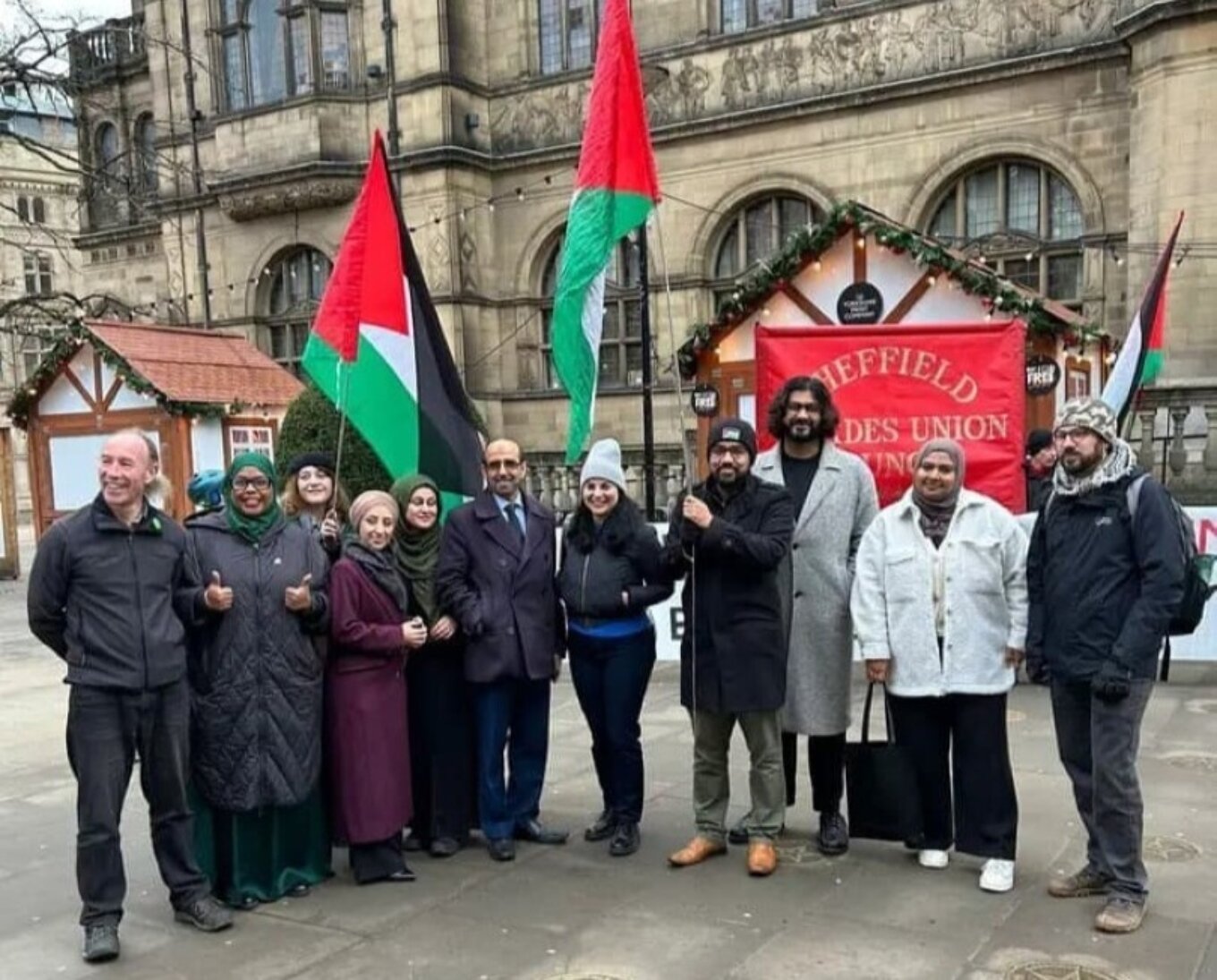 A photo of eleven people standing side by side outside Sheffield Town Hall and smiling at the camera, with two holding big Palestinian flags