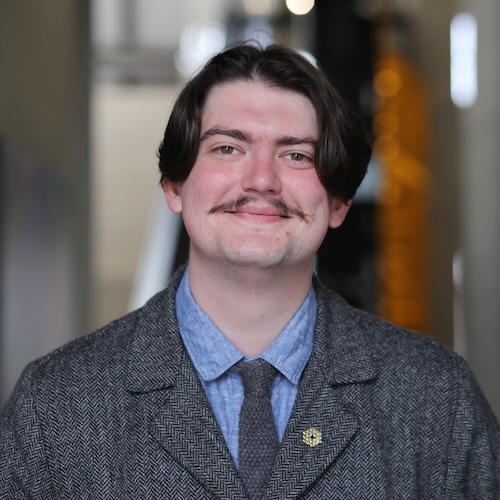 Smiling man with long, thin moustache wearing a suit jacket and tie.