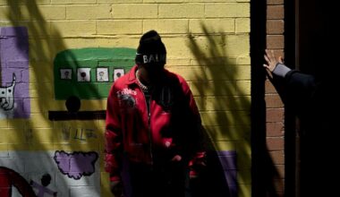 T-Man, a Black teen wearing a red coat, stands in front of a yellow wall that has a mural painted on it. His face is not visible.