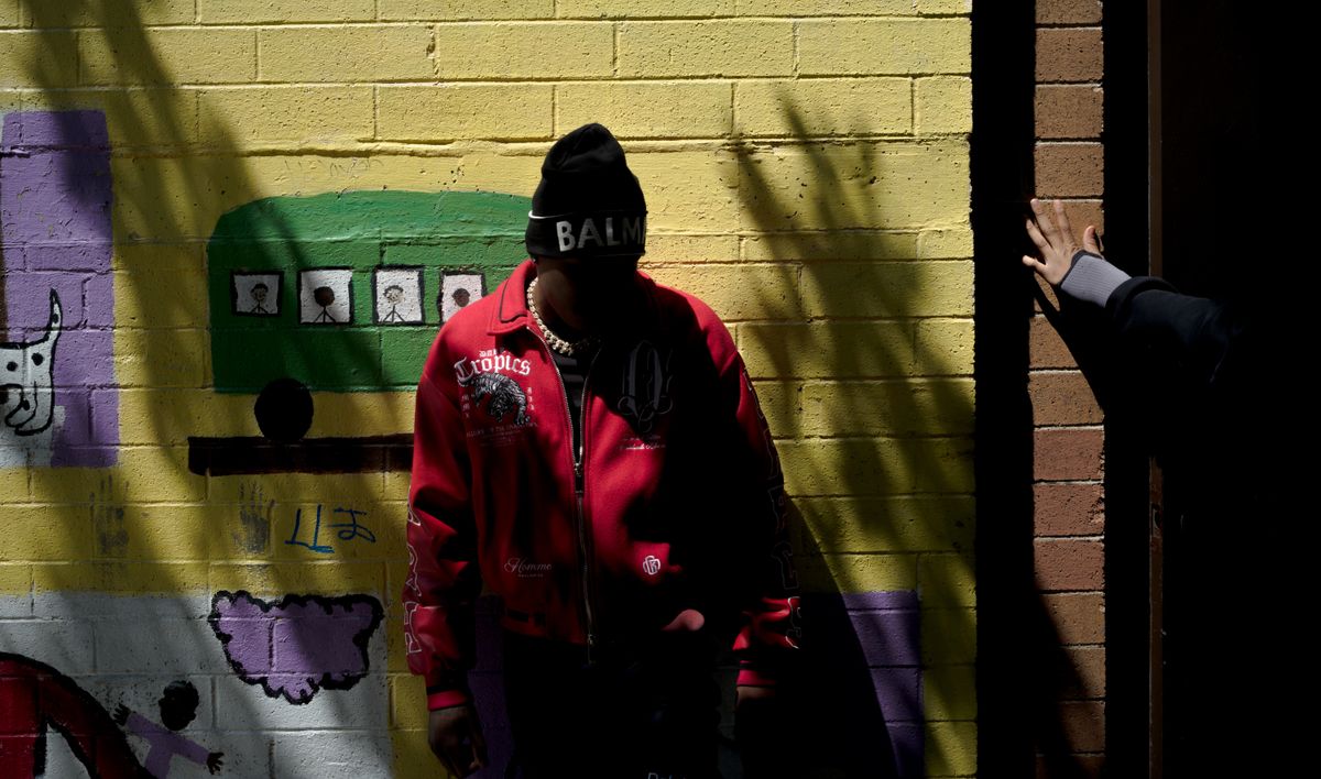 T-Man, a Black teen wearing a red coat, stands in front of a yellow wall that has a mural painted on it. His face is not visible.