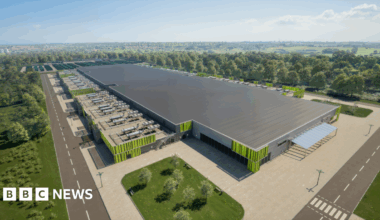 An artist's impression of the proposed new green AI energy centre in Ravenscraig - a large building with a grey roof and  green design on the walls. A car park is part of the site with entry roads around it.