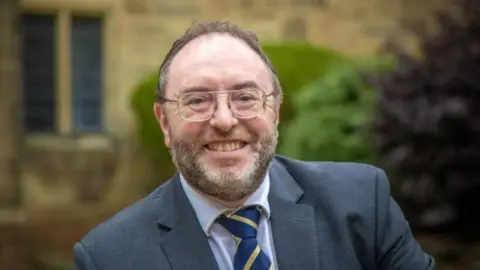 Bangor University Edmund Burke smiles at the camera. He has short brown hair and a beard slightly greying. He is wearing square-framed glasses and a grey suit jacket. A white shirt and a blue and yellow stripe tie. Behind him a blurred background of a historic yellow brick building.