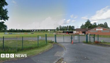 Google street view of the West Bank Bears ground in Widnes. There is a club house with a pitch in the background, along with advertisement boards. There is a green wire fence and a gate in the foreground.