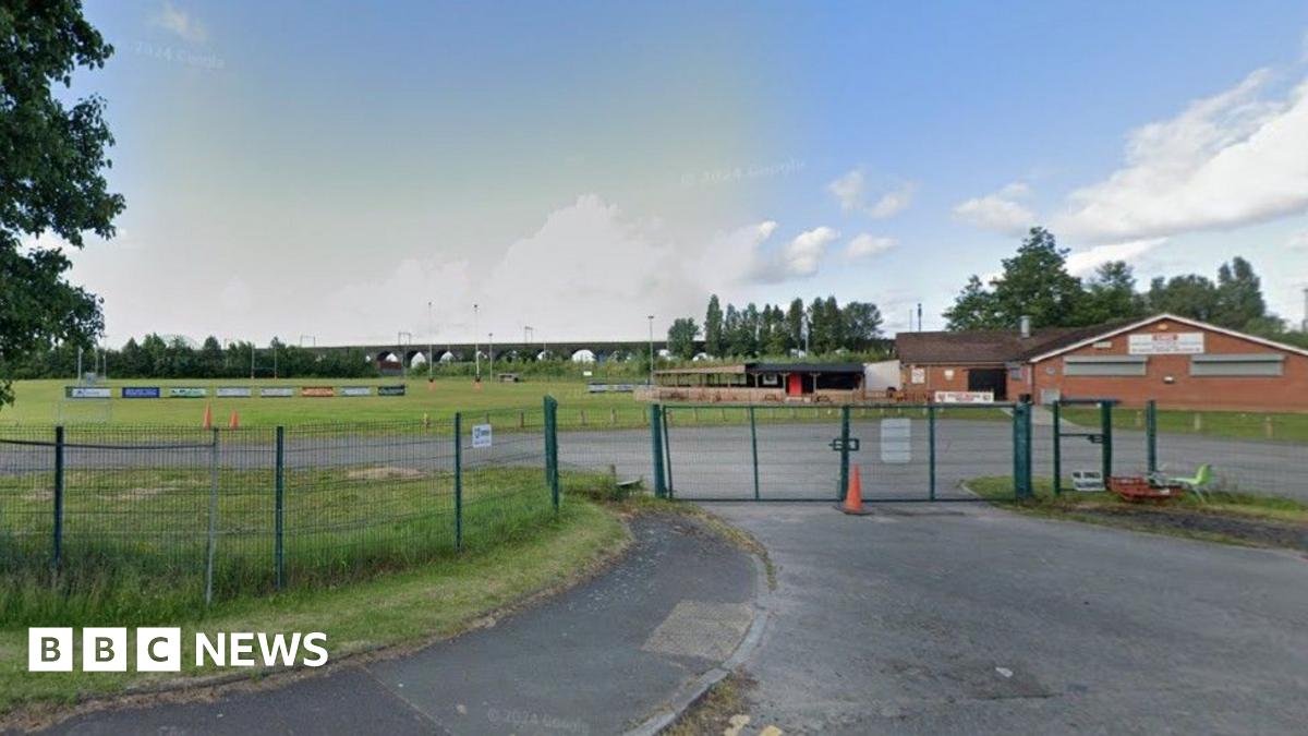 Google street view of the West Bank Bears ground in Widnes. There is a club house with a pitch in the background, along with advertisement boards. There is a green wire fence and a gate in the foreground.