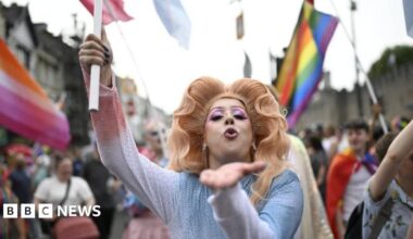 A person with a large wig and pink eyeliner blows a kiss at the camera