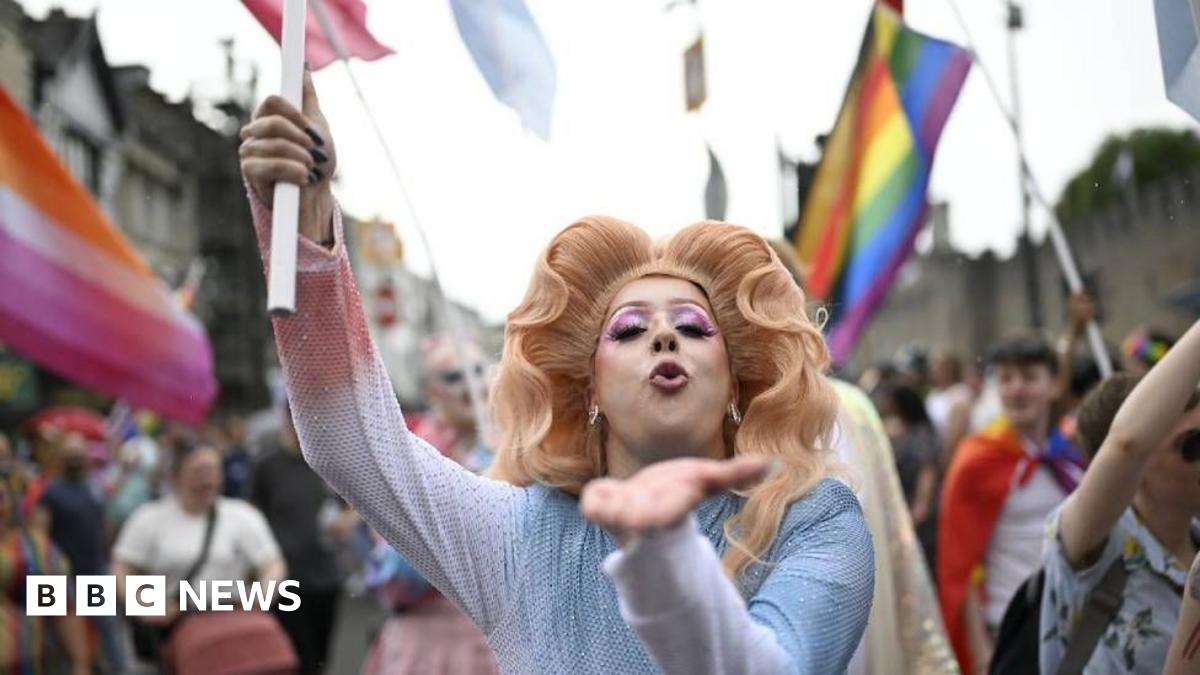 A person with a large wig and pink eyeliner blows a kiss at the camera