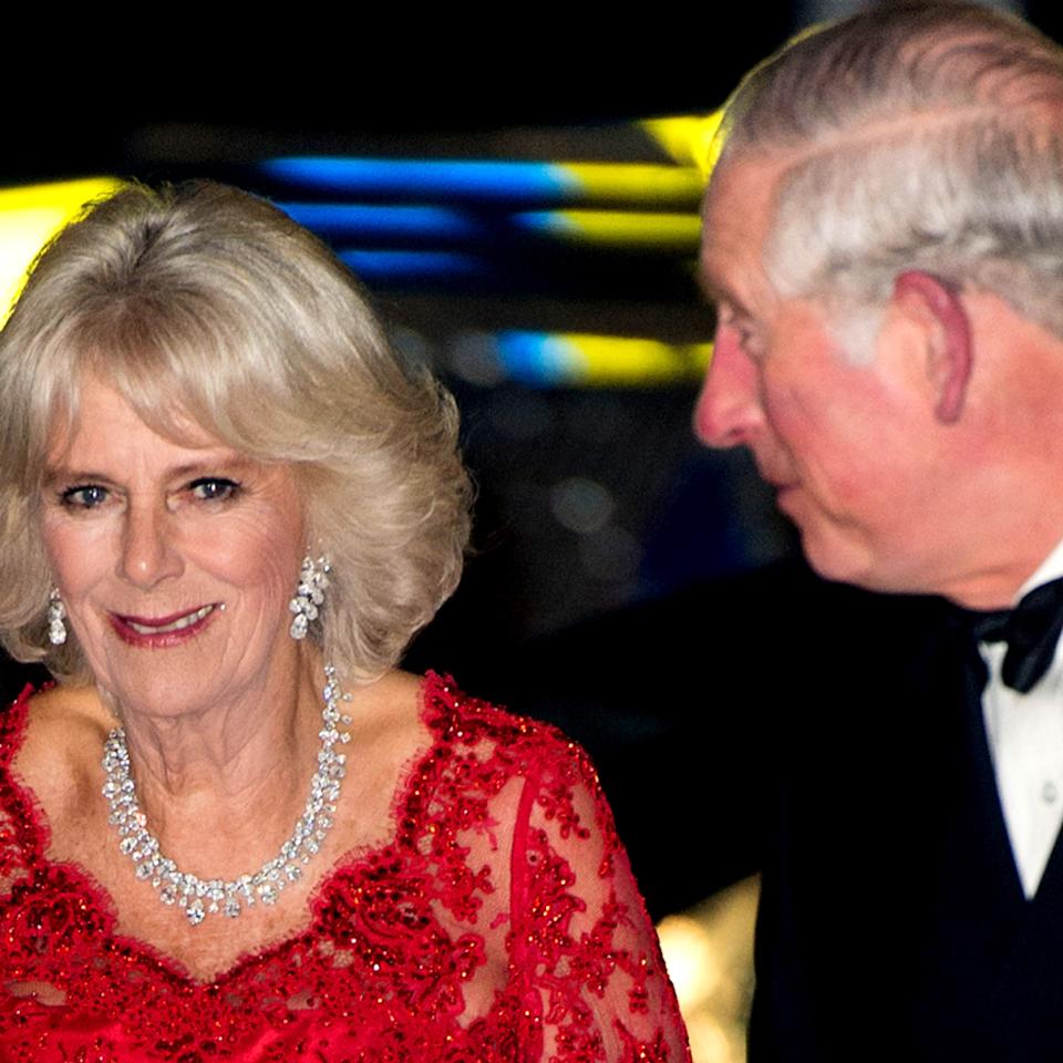  Queen Camilla wearing a red dress and diamonds next to King Charles in a tux. 