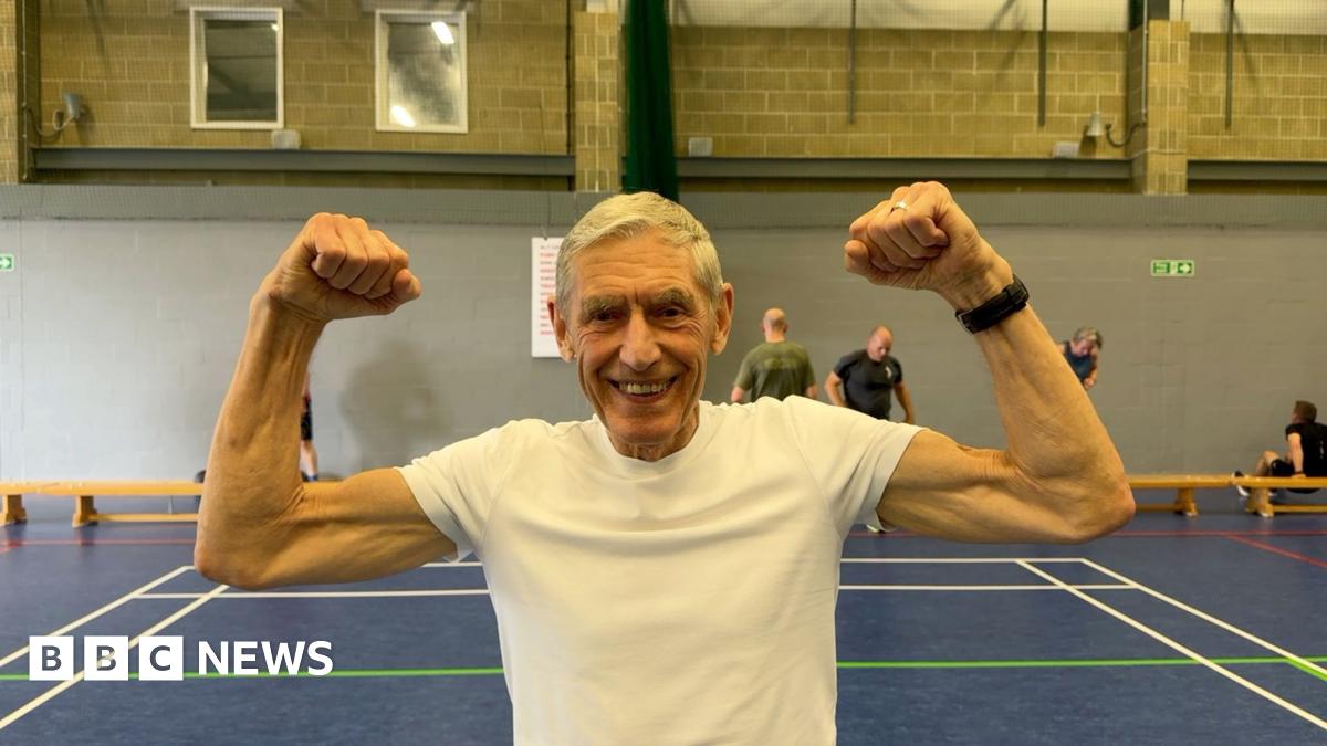 A man is stood in a gymnasium flexing his muscles. He is wearing a white t-shirt and there are people training behind him.