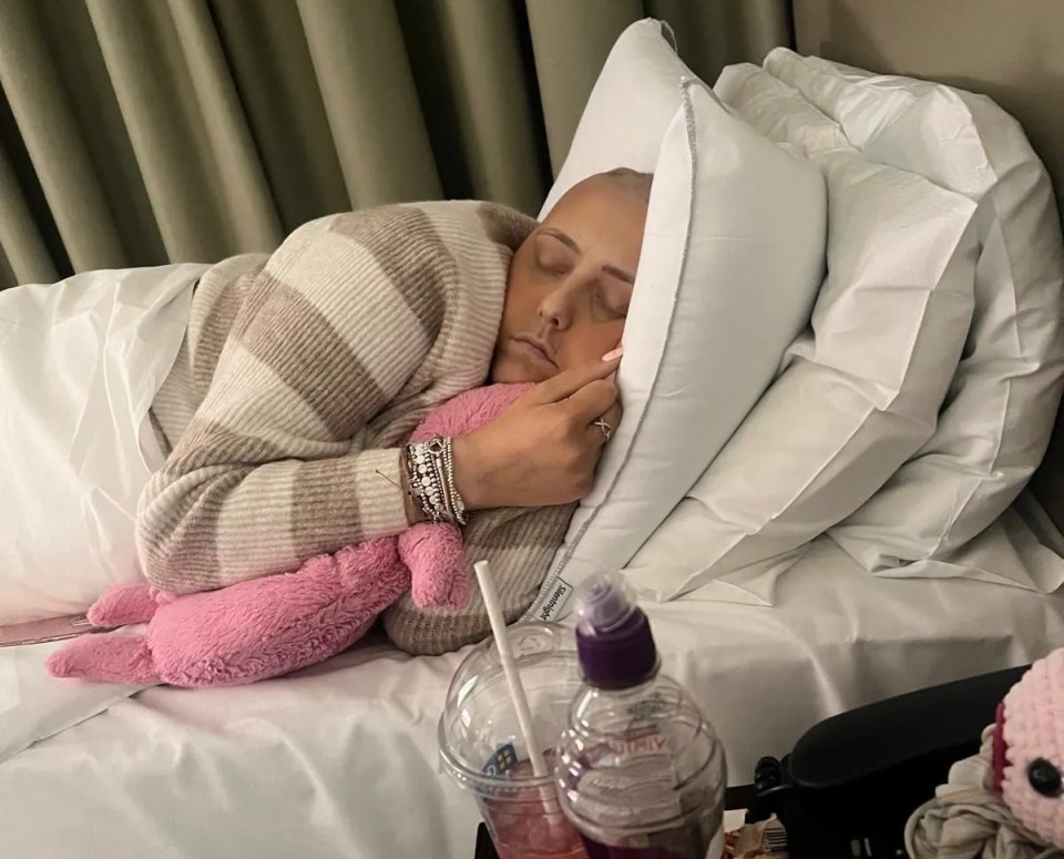 A young woman with no hair sleeps in a hospital bed, holding a pink stuffed animal.
