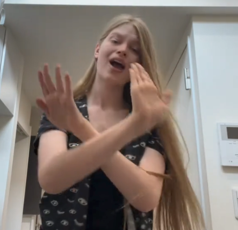 Person with long hair and patterned shirt making expressive hand gestures in a hallway