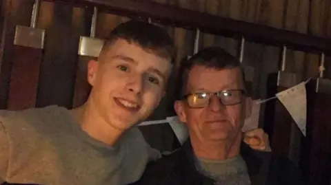 Tom McGrath Matthews McGrath has short brown hair, his smiling and wearing a grey sweatshirt with his arm over his father. Tom McGrath has short brown hair, he is wearing black square frame glasses, a grey jumper and a black jacket. Behind them is white bunting tied to a set of bannisters. 