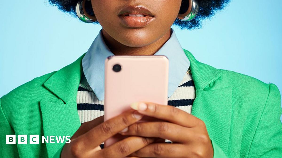 A woman looking at her smartphone. she is wearing a green jacket, blue shirt and blue and white stripe top and is in front of a plain blue background