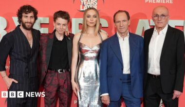 Four actors and Danny Boyle pose in a row, standing against a promotional red board featuring the title of the film 28 Years Later.