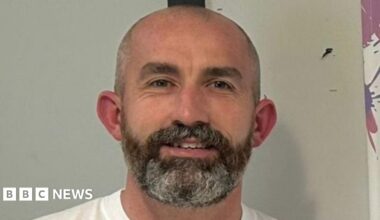 Gerard Donaghy is standing in front of a white wall, wearing a white t-shirt and is smiling at the camera. He has a bald head and a dark beard with grey hair through it.