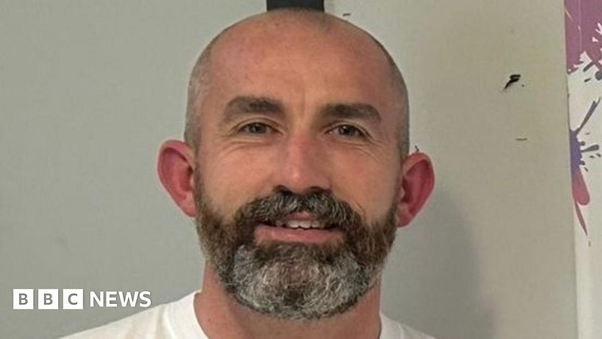 Gerard Donaghy is standing in front of a white wall, wearing a white t-shirt and is smiling at the camera. He has a bald head and a dark beard with grey hair through it.