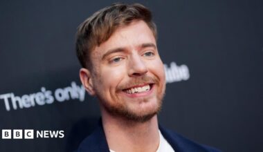 MrBeast pictured in New York in May 2025. The creator has blue eyes, short brown hair and a short beard. He smiles, wearing a dark blue blazer over a white T-shirt while posing for photographers in front of black hoarding.