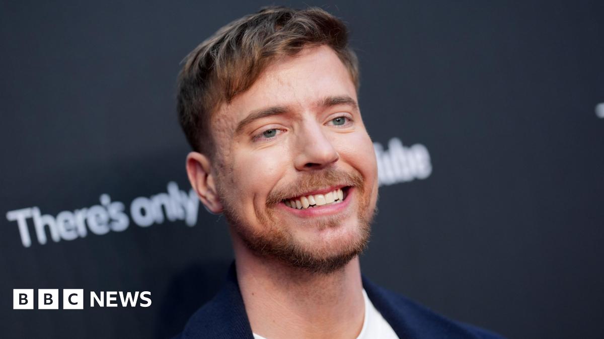MrBeast pictured in New York in May 2025. The creator has blue eyes, short brown hair and a short beard. He smiles, wearing a dark blue blazer over a white T-shirt while posing for photographers in front of black hoarding.