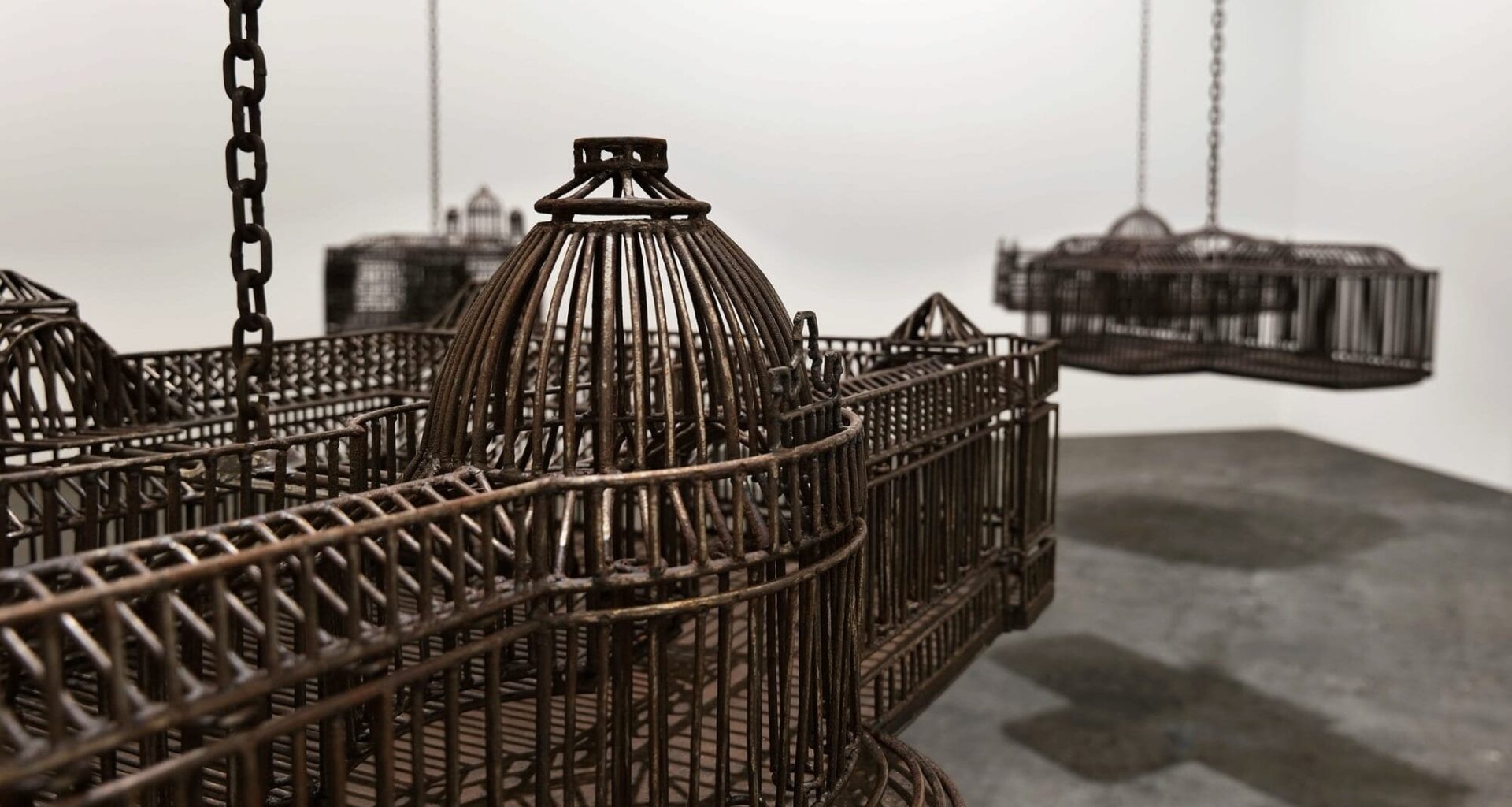 detail of an installation in a white-walled gallery space of sculptures suspended from the ceiling by chains, each one modeled after a colonial, institutional building