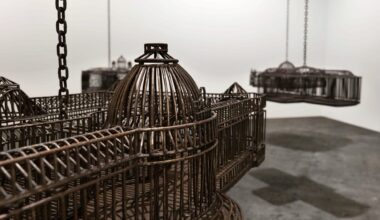 detail of an installation in a white-walled gallery space of sculptures suspended from the ceiling by chains, each one modeled after a colonial, institutional building