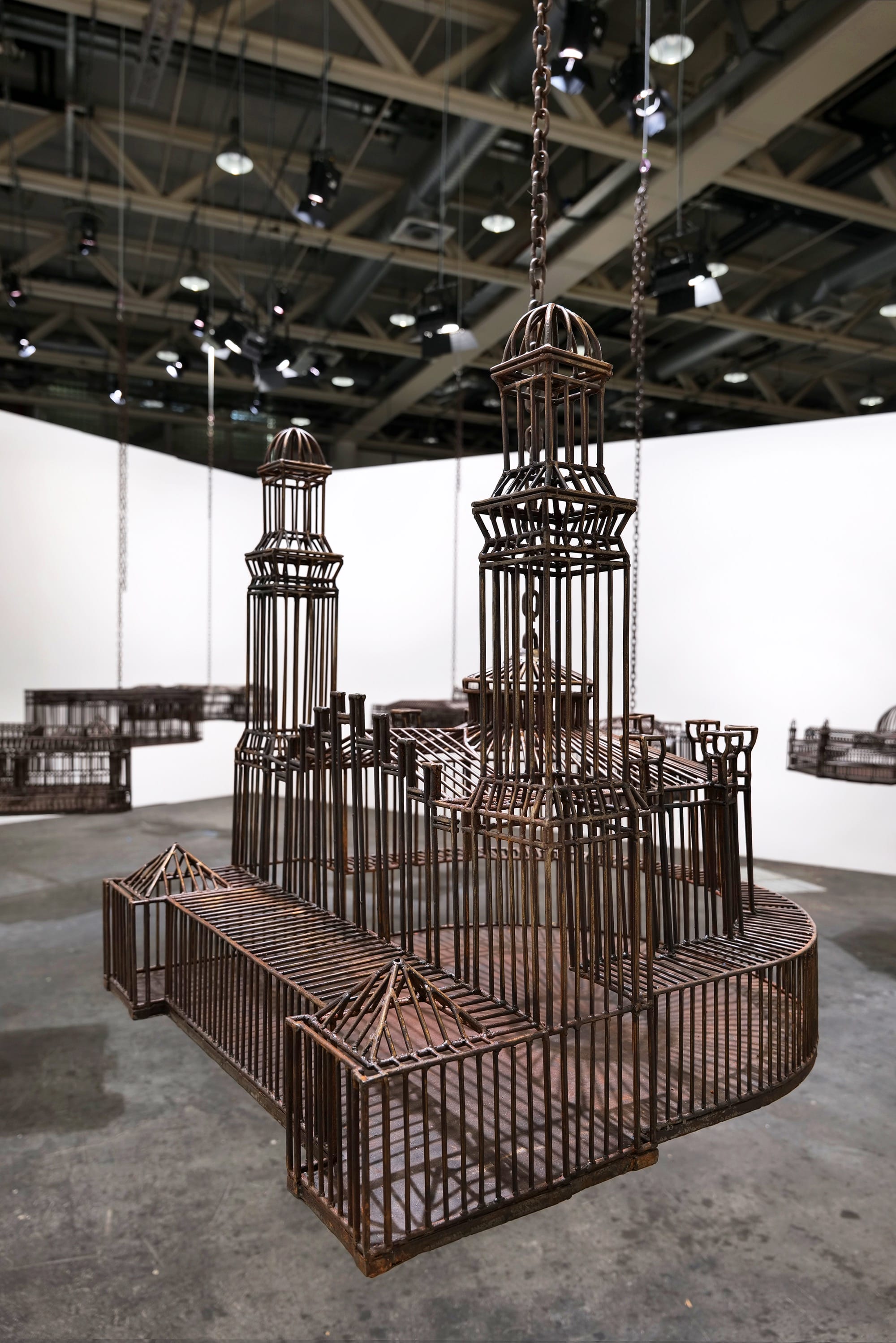 detail of an installation in a white-walled gallery space of sculptures suspended from the ceiling by chains, each one modeled after a colonial, institutional building