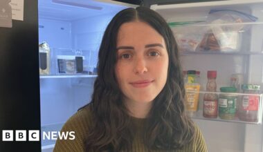 A lady with shoulder length brown hair smiles as she wears a green jumper. She is standing in front of an open fridge.