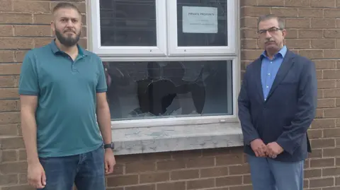 Kashif Akram and Raied Al-Wazzan standing beside the window that was brokem in the attack on the Belfast Islamic Centre.  Mr Akram has short brown hair and a beard and is wearing a green polo shirt and blue jeans.   Mr Al-Wazzan has short grey hair and a grey moustache.  He is wearing glasses, a blue shirt, a navy suit jacket and grey trousers. 