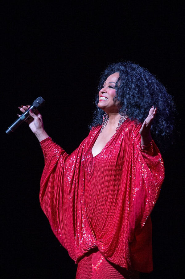 Diana Ross in a sparkly red dress holding a microphone, smiling and holding her arms out