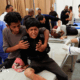 A boy sitting on a bed crises, while a man sat beside him hugs him and a woman stands beside him