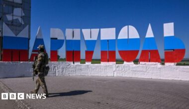 A Russian soldier stands in the ruins of Mariupol's theatre in spring 2022, taking a photo out of a destroyed window using his phone