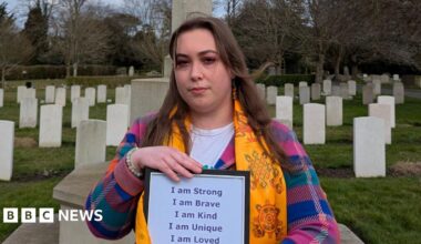 Claire wearing a colourful chequered jumper and a yellow scarf while standing in front of graves in a graveyard. She has long brown hair and is holding a piece of paper that says she is strong, brave, kind, unique and loved.