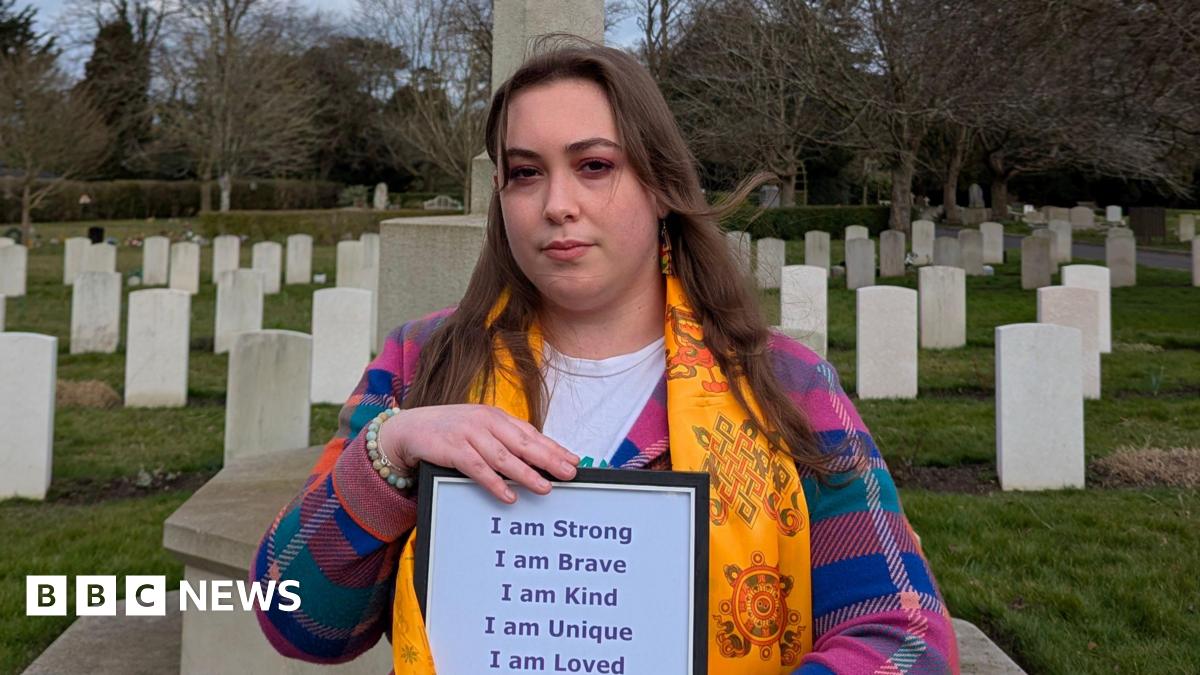 Claire wearing a colourful chequered jumper and a yellow scarf while standing in front of graves in a graveyard. She has long brown hair and is holding a piece of paper that says she is strong, brave, kind, unique and loved.