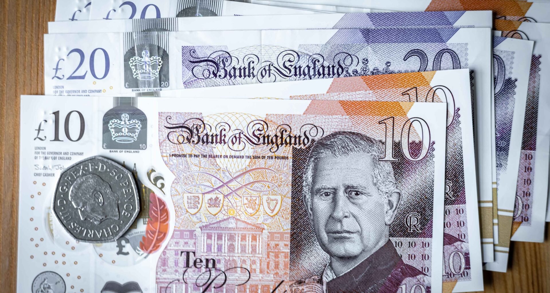 Ten pound sterling note on top of other notes, a 50 pence piece is on top of the ten pound note. The note and coin carry the image of the King.