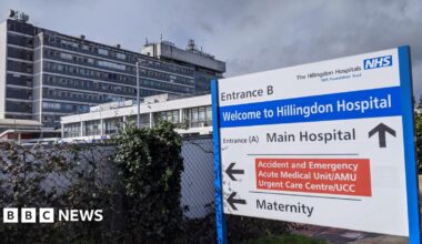 A general view of Entrance b of Hillingdon Hospital. The sign is blue, white and red. In the background, there was high buildings with a dark cloud and some leaves.