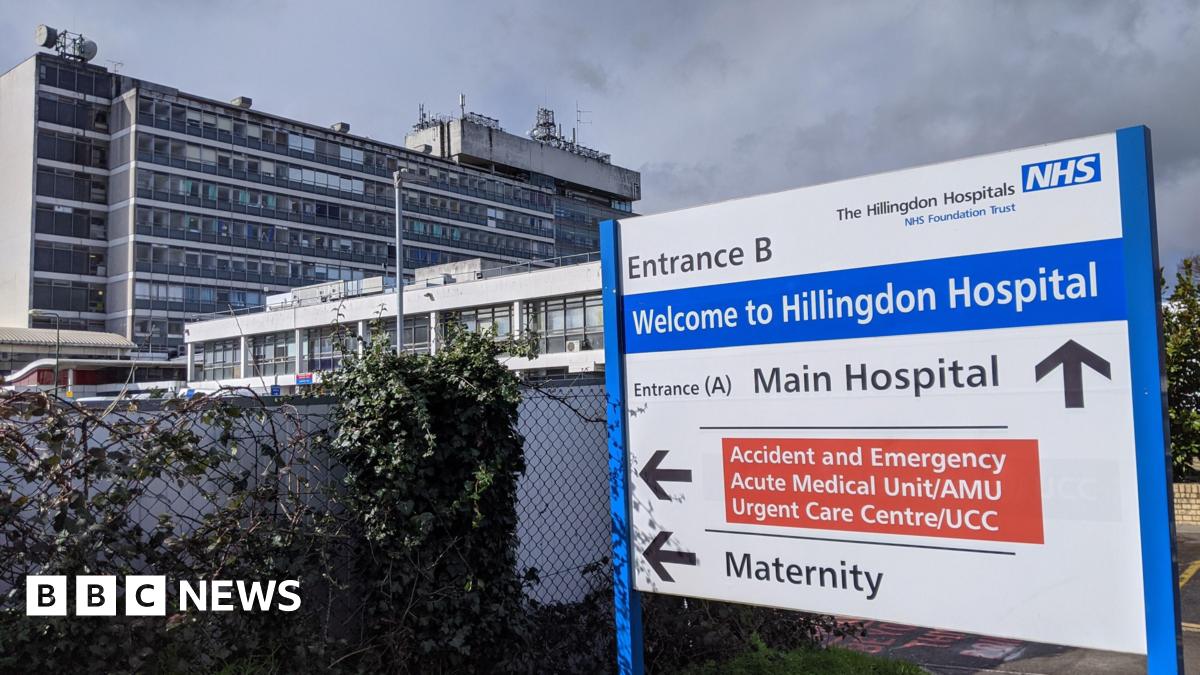 A general view of Entrance b of Hillingdon Hospital. The sign is blue, white and red. In the background, there was high buildings with a dark cloud and some leaves.