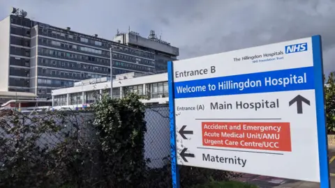 Anahita Hossein-Pour A general view of Entrance b of Hillingdon Hospital. The sign is blue, white and red. In the background, there was high buildings with a dark cloud and some leaves.
