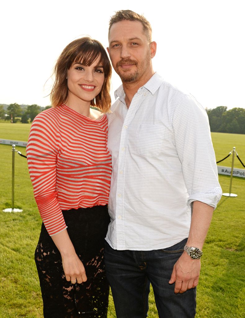 Charlotte Riley wears a red stripey top with black skirt and Tom Hardy stands next to her wearing a white shirt with jeans with polo pitch at Coworth Park, Sunningdale, in the background
