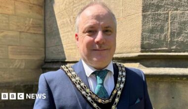 A balding man with short grey hair on the back and sides of his head, wearing a gold chain, a blue suit jacket, green tie, white shirt, and double chain, stands outside of a church.