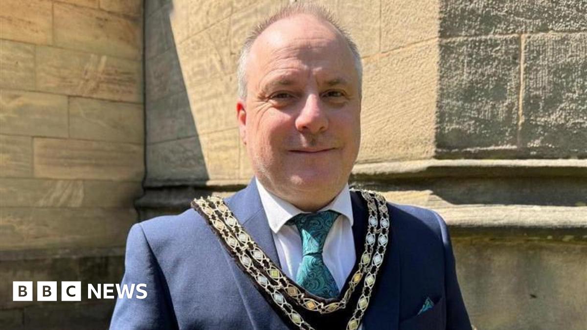 A balding man with short grey hair on the back and sides of his head, wearing a gold chain, a blue suit jacket, green tie, white shirt, and double chain, stands outside of a church.
