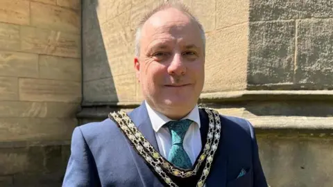 Adam Parkes A balding man with short grey hair on the back and sides of his head, wearing a gold chain, a blue suit jacket, green tie, white shirt, and double chain, stands outside of a church. 