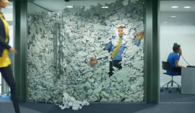 A businessman is trapped in his glass office by a surplus of discarded ideas on paper . His colleague in the next office is working more efficiently and is oblivious to him being trapped , as is a passing female office worker