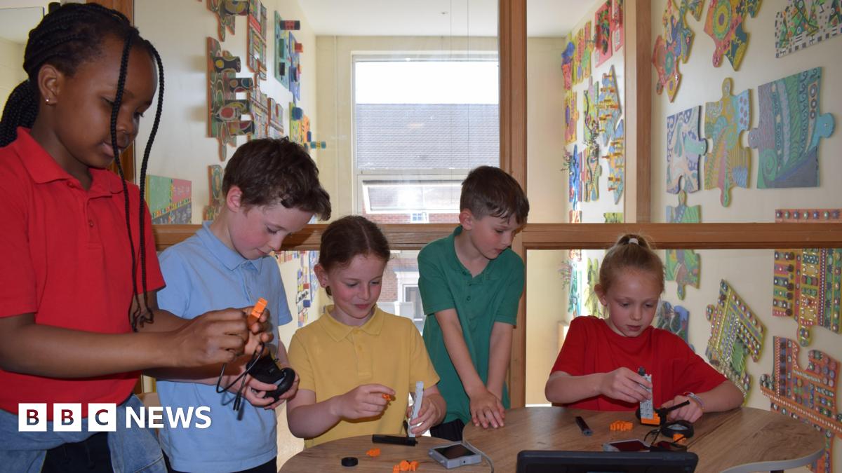 Swindon school children building with Lego style blocks, all wearing bright air tech style tops. Two children are wearing red, one is wearing yellow, one is in blue and one in green. They are gathered around a central table all focussed on what they are building. In the background, there is a wall with big puzzle piece as art on it.