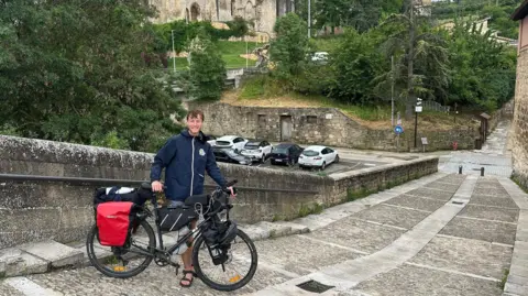Ollie White Ollie wears a dark blue rain coat and poses with his black bike on a steep cobbled ramp. There are a few cars and a large stone building behind him. 