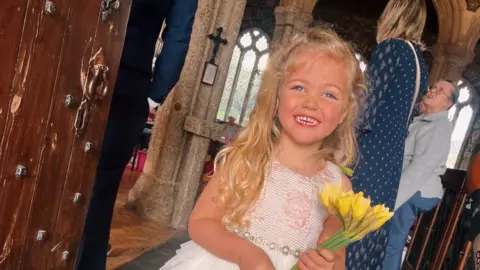Sharon Hart Willow has blonde hair and blue eyes and wears a white dress while holding yellow flowers as she smiles toward the camera in a church 